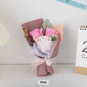 K&ouml;nnte beinhalten: Ein kleiner Strau&szlig; aus rosa und wei&szlig;en Rosen, verpackt in mauvefarbenem Papier und mit einem Band mit der Aufschrift "Love is" gebunden. Der Strau&szlig; enth&auml;lt Zweige mit Gr&uuml;n und wird auf einer wei&szlig;en Oberfl&auml;che pr&auml;sentiert.