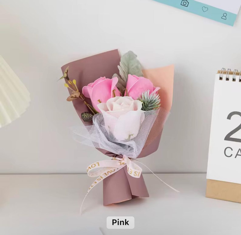 K&ouml;nnte beinhalten: Ein kleiner Strau&szlig; aus rosa und wei&szlig;en Rosen, verpackt in mauvefarbenem Papier und mit einem Band mit der Aufschrift "Love is" gebunden. Der Strau&szlig; enth&auml;lt Zweige mit Gr&uuml;n und wird auf einer wei&szlig;en Oberfl&auml;che pr&auml;sentiert.