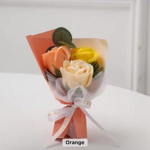 K&ouml;nnte beinhalten: Ein kleines Bouquet aus Seifenrosen in Orange-, Gelb- und Cremet&ouml;nen, verpackt in pfirsichfarbenem Papier und mit einem wei&szlig;en Band gebunden. Das Bouquet enth&auml;lt ein kleines gr&uuml;nes Blatt und ist mit "Orange" beschriftet.