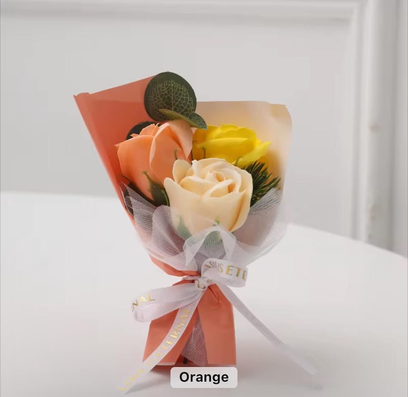 K&ouml;nnte beinhalten: Ein kleines Bouquet aus Seifenrosen in Orange-, Gelb- und Cremet&ouml;nen, verpackt in pfirsichfarbenem Papier und mit einem wei&szlig;en Band gebunden. Das Bouquet enth&auml;lt ein kleines gr&uuml;nes Blatt und ist mit "Orange" beschriftet.