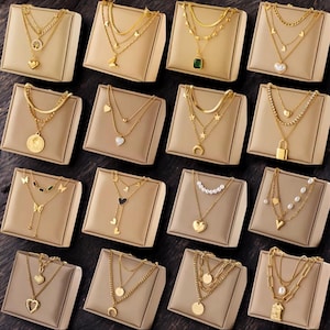 May include: A collection of gold-colored necklaces displayed on beige stands. The necklaces feature various pendants, including hearts, coins, and geometric shapes. Some necklaces have multiple layers, and others include gemstones and pearls.