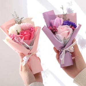 K&ouml;nnte beinhalten: Zwei kleine Str&auml;u&szlig;e aus Seifenblumen, einer in Rosa und der andere in Lila. Jeder Strau&szlig; ist in farbiges Papier gewickelt und mit einem Band gebunden. Die Str&auml;u&szlig;e enthalten Rosen, Nelken und andere kleine Blumen.