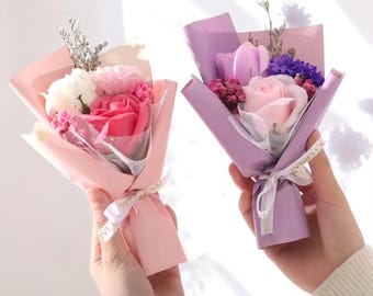 May include: Two small bouquets of soap flowers, one in pink and the other in purple. Each bouquet is wrapped in colored paper and tied with a ribbon. The bouquets contain roses, carnations, and other small flowers.