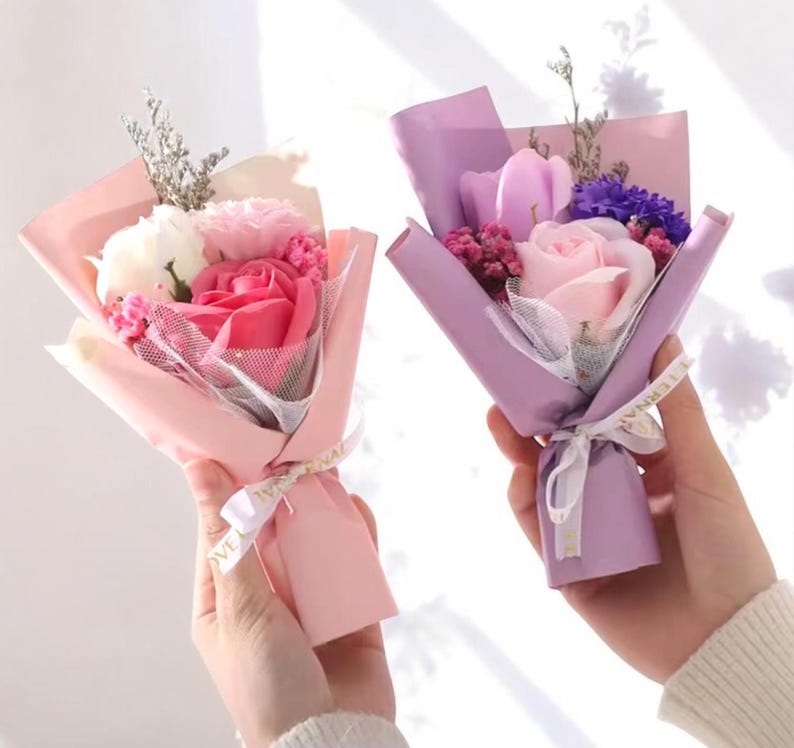 K&ouml;nnte beinhalten: Zwei kleine Str&auml;u&szlig;e aus Seifenblumen, einer in Rosa und der andere in Lila. Jeder Strau&szlig; ist in farbiges Papier gewickelt und mit einem Band gebunden. Die Str&auml;u&szlig;e enthalten Rosen, Nelken und andere kleine Blumen.