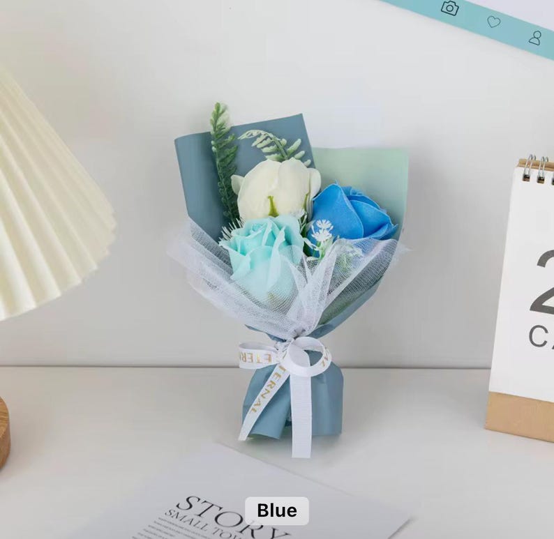 K&ouml;nnte beinhalten: Ein kleines Bouquet aus k&uuml;nstlichen Rosen in Blau-, Wei&szlig;- und Hellblaut&ouml;nen, verpackt in blauem Papier und wei&szlig;em T&uuml;ll. Das Bouquet ist mit einem wei&szlig;en Band mit dem Aufdruck "Eternal" gebunden. Das Wort "Blue" ist auf einem wei&szlig;en Etikett aufgedruckt.