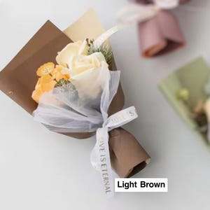 K&ouml;nnte beinhalten: Ein kleiner Strau&szlig; aus cremefarbenen und orangefarbenen Blumen, verpackt in braunem und beigem Papier, gebunden mit einem wei&szlig;en Band mit der Aufschrift "LOVE IS ETERNAL". Der Strau&szlig; ist teilweise mit wei&szlig;em Netz bedeckt.