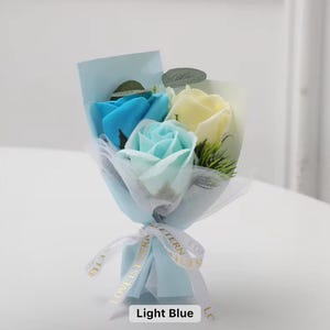K&ouml;nnte beinhalten: Ein kleines Bouquet aus Seifenrosen in Blau- und Gelbt&ouml;nen, verpackt in hellblauem Papier und T&uuml;ll. Der Strau&szlig; ist mit einem wei&szlig;en Band gebunden, auf dem "LOVE IS ETERNAL" steht. Der Text "Light Blue" befindet sich unten.