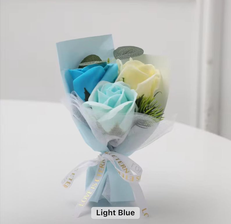 K&ouml;nnte beinhalten: Ein kleines Bouquet aus Seifenrosen in Blau- und Gelbt&ouml;nen, verpackt in hellblauem Papier und T&uuml;ll. Der Strau&szlig; ist mit einem wei&szlig;en Band gebunden, auf dem "LOVE IS ETERNAL" steht. Der Text "Light Blue" befindet sich unten.
