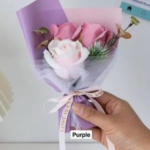 K&ouml;nnte beinhalten: Ein kleiner Strau&szlig; aus rosa und wei&szlig;en Rosen, verpackt in lila und rosa Papier, gebunden mit einem wei&szlig;en Band mit der Aufschrift "LOVE IS ET". Das Wort "Purple" ist unten aufgedruckt.