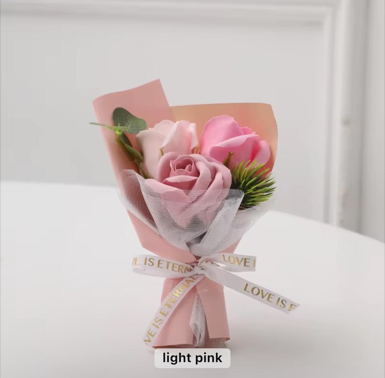 K&ouml;nnte beinhalten: Ein kleiner Strau&szlig; hellrosa Rosen, eingewickelt in rosa Papier und wei&szlig;em Netz. Der Strau&szlig; ist mit einem Band gebunden, auf dem "LOVE IS ETERNAL" steht. Der Text "light pink" steht unten.