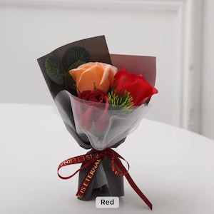 K&ouml;nnte beinhalten: Ein kleines Bouquet aus Seifenrosen in Rot- und Oranget&ouml;nen, verpackt in schwarzem und braunem Papier mit einem wei&szlig;en Netz&uuml;berzug. Das Bouquet ist mit einem roten Band mit der Aufschrift "IS ETERNAL" gebunden. Das Wort "Red" ist auf einem kleinen wei&szlig;en Rechteck aufgedruckt.