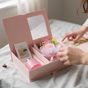 May include: A pink gift box with a mirror inside, containing a perfume bottle, a small card, and a decorative flower arrangement. The box is open, revealing the contents. The overall theme is a gift set.