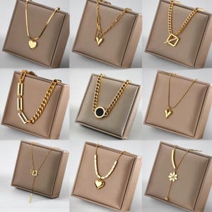 May include: Nine gold-colored necklaces displayed on neutral-toned pedestals. The necklaces feature various designs, including heart pendants, geometric shapes, and chain links. The jewelry is presented in a studio setting, highlighting the different styles and designs.