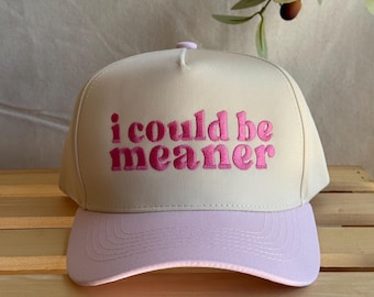 May include: A beige baseball cap with a light purple brim. The phrase "i could be meaner" is embroidered in pink across the front of the cap. The cap is sitting on a wooden surface.