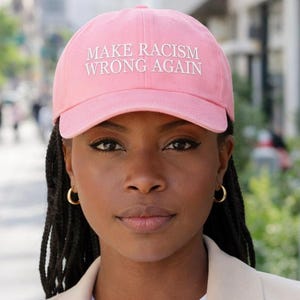 May include: A pink baseball cap with the text "MAKE RACISM WRONG AGAIN" embroidered in white. The cap is worn by a person wearing a white shirt and a beige blazer.