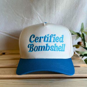 May include: A white and blue trucker hat with the text "Certified Bombshell" embroidered in blue. The hat has a curved brim and a mesh back, suitable for casual wear.