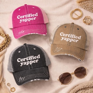 May include: Three distressed trucker hats in pink, tan, and black, each with the text "Certified Yapper" in white. The hats have mesh backs and distressed visors. Sunglasses and jewelry are also visible.