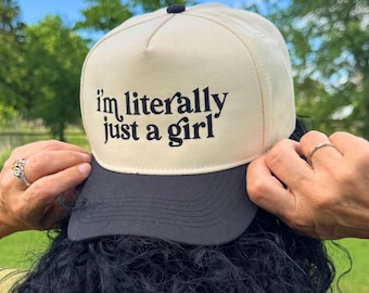 May include: A beige and black baseball cap with the text "I'm literally just a girl" in black script. The cap has a black brim and a black button on top. The cap is being held by a person.