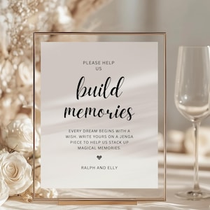 Puede incluir: Un letrero enmarcado transparente con el texto "PLEASE HELP US build memories". El letrero incluye el texto "EVERY DREAM BEGINS WITH A WISH. WRITE YOURS ON A JENGA PIECE TO HELP US STACK UP MAGICAL MEMORIES. RALPH AND ELLY".
