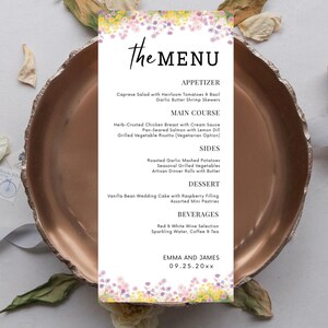 Pode incluir: Um cartão de menu de casamento com detalhes florais, apresentando o texto "the MENU" em uma escrita elegante. O menu lista aperitivos, pratos principais, acompanhamentos, sobremesas e bebidas. Os nomes "Emma e James" e uma data também estão incluídos.