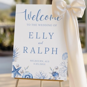 May include: A white wedding welcome sign with blue text and seashell illustrations. The sign reads "Welcome" and announces the wedding of Elly and Ralph in Melbourne, AUS. A large cream bow adorns the side, and the sign is on a gold easel.