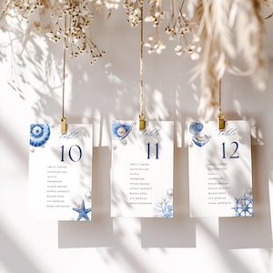 Puede incluir: Tres tarjetas colgantes con números de mesa con diseños náuticos. Cada tarjeta presenta una concha azul acuarela, detalles de perlas y el número de mesa en azul marino. Las tarjetas están suspendidas por clips dorados.