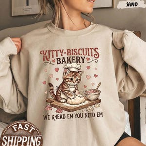 May include: A sand-colored sweatshirt featuring a cartoon kitten chef kneading dough. The text "KITTY BISCUITS BAKERY" arches above the kitten, with hearts and the phrase "WE KNEAD EM YOU NEED EM" below. The design includes a wooden cutting board, bowl, and pink pastries.