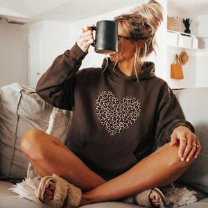 May include: A brown hoodie with a large leopard print heart design. The hoodie has a front pocket and a drawstring hood. The person is holding a black mug. The person is wearing fuzzy slippers.
