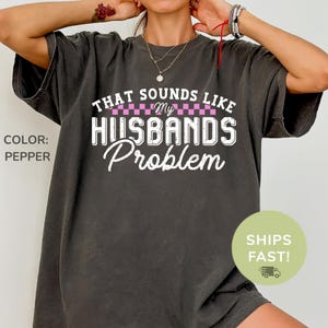 May include: Pepper-colored t-shirt with the text "THAT SOUNDS LIKE MY HUSBANDS Problem" in white and pink lettering. The shirt has a crew neck and short sleeves. The color is listed as pepper.