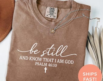 May include: Espresso-colored t-shirt with the words "be still" and "AND KNOW THAT I AM GOD PSALM 46:10" printed in white. A silver necklace with a pendant rests on the shirt. The shirt is a Comfort Colors brand.