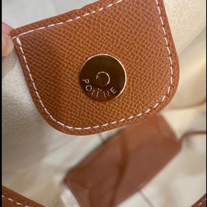 Puede incluir: Primer plano de un bolso de mano de cuero marr&oacute;n con un cierre magn&eacute;tico dorado. El bolso presenta costuras blancas y una superficie texturizada. El interior es de color beige claro.