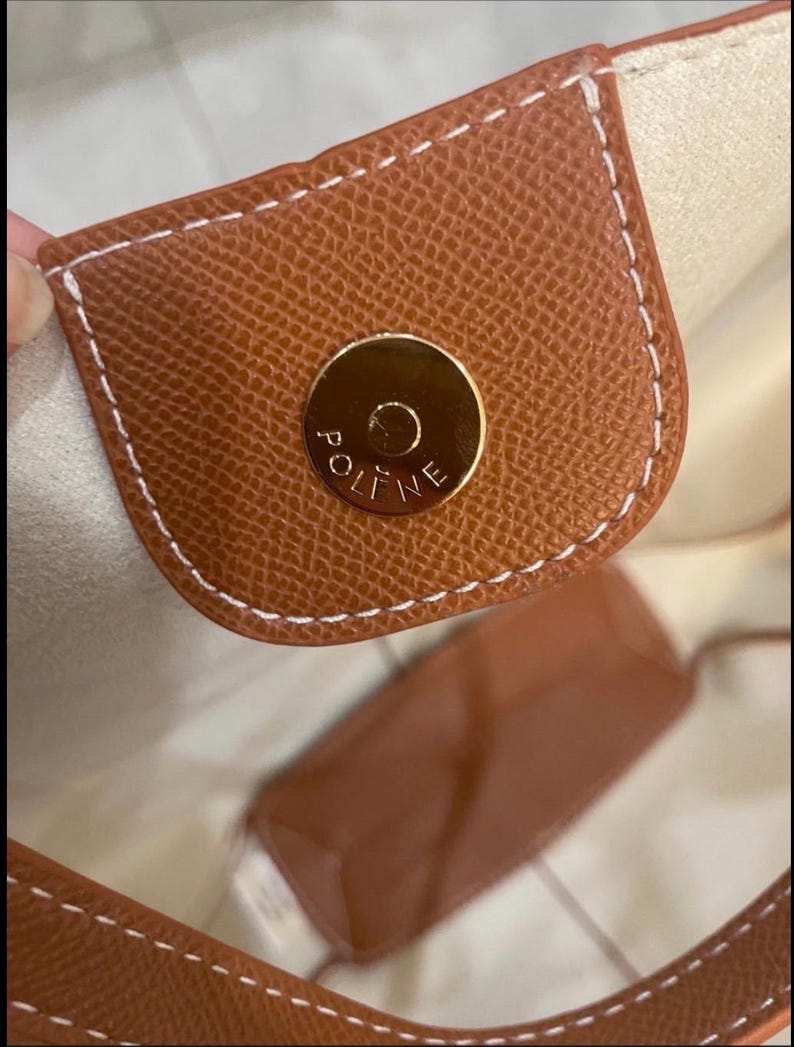 Puede incluir: Primer plano de un bolso de mano de cuero marr&oacute;n con un cierre magn&eacute;tico dorado. El bolso presenta costuras blancas y una superficie texturizada. El interior es de color beige claro.