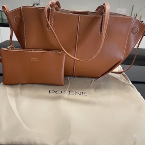 Puede incluir: Un bolso tote de cuero marr&oacute;n y una peque&ntilde;a bolsa a juego. El tote tiene costuras blancas y asas. La bolsa tiene cierre de cremallera. El conjunto se muestra en una bolsa guardapolvo beige con la marca "POLENE" impresa.