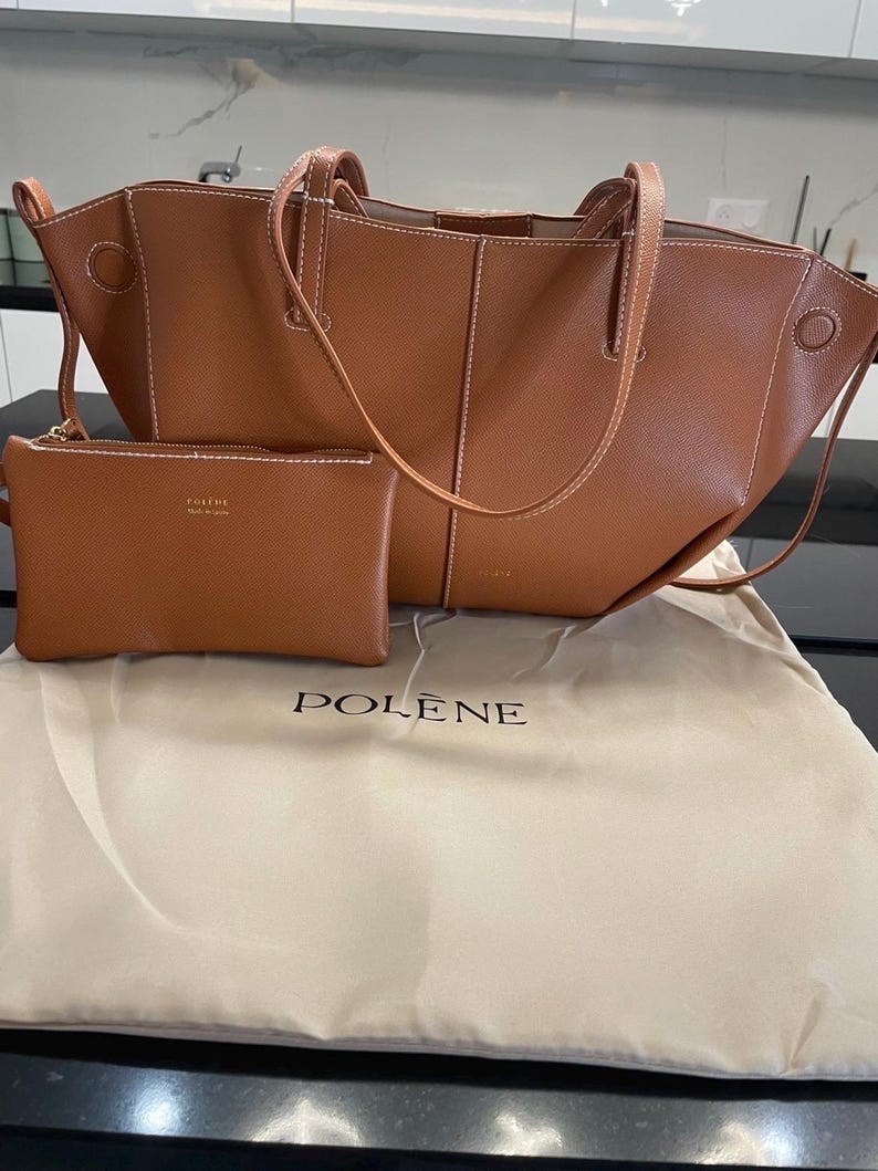 Puede incluir: Un bolso tote de cuero marr&oacute;n y una peque&ntilde;a bolsa a juego. El tote tiene costuras blancas y asas. La bolsa tiene cierre de cremallera. El conjunto se muestra en una bolsa guardapolvo beige con la marca "POLENE" impresa.