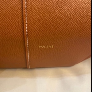 Puede incluir: Primer plano de un bolso de mano de cuero marr&oacute;n con una superficie texturizada. Una costura blanca recorre verticalmente el centro. La palabra "POL&Egrave;NE" est&aacute; impresa en letras doradas en el bolso.