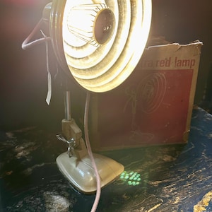 May include: A vintage, white and black infrared lamp with a metal cage around the bulb. The lamp is on a table and is plugged in. The lamp is in its original box, which says "Infrared Lamp" on it.