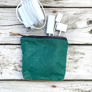 May include: A green waxed canvas zippered pouch with a brown leather zipper pull. The pouch is sitting on a white wooden surface with white cords and white adapters.
