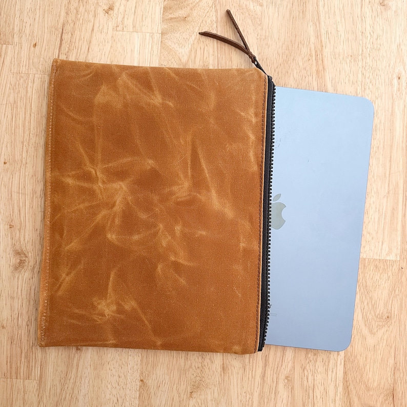 May include: A rectangular tan waxed canvas sleeve with a black zipper closure and a leather pull tab. A light blue tablet with a silver apple logo is partially inserted into the sleeve, revealing a portion of the device's smooth metallic surface.