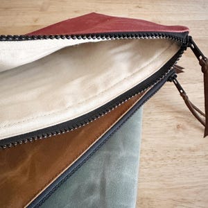 May include: A set of three zippered pouches featuring a deep red, a tan, and a sage green exterior. Each pouch has a black zipper closure with a brown leather pull tab and a plain off-white fabric lining visible inside the open red pouch.