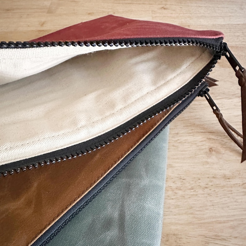 May include: A set of three zippered pouches featuring a deep red, a tan, and a sage green exterior. Each pouch has a black zipper closure with a brown leather pull tab and a plain off-white fabric lining visible inside the open red pouch.