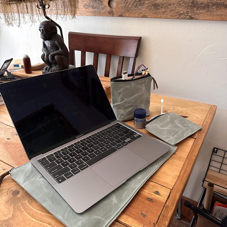 May include: A silver laptop sits on a sage green waxed canvas sleeve. Next to it are a matching sage green zippered pouch containing cables, a small sage green wallet, and a blue cylindrical container with white text on a wooden surface.