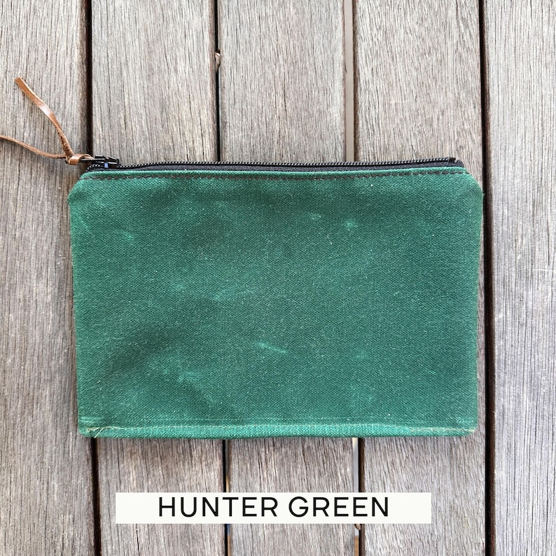 May include: A rectangular hunter green zippered pouch featuring a black zipper closure and a thin brown leather pull tab. The pouch is constructed from a textured fabric and includes the text HUNTER GREEN printed in black on a white rectangular label.