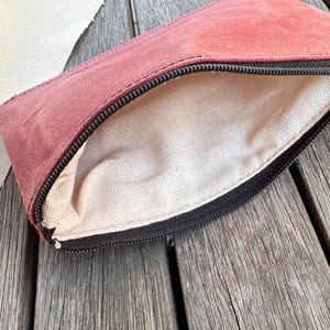 May include: A rectangular zippered pouch featuring a dusty rose exterior made of a textured fabric. The interior is lined with a plain, off-white canvas material. A black zipper runs along the top edge, providing a contrast to the muted pink exterior.