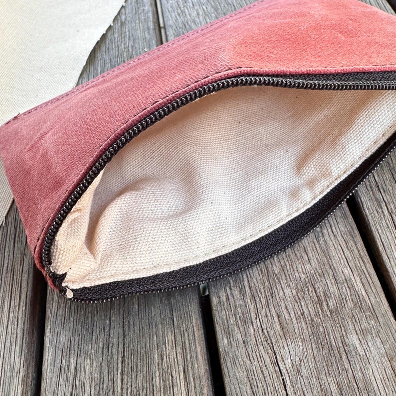 May include: A rectangular zippered pouch featuring a dusty rose exterior made of a textured fabric. The interior is lined with a plain, off-white canvas material. A black zipper runs along the top edge, providing a contrast to the muted pink exterior.