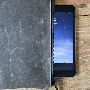 May include: A dark olive green zippered pouch with a textured, weathered finish and a black zipper closure. A small tablet device with a visible screen showing a starry sky background and the time is partially inserted into the open side of the pouch.
