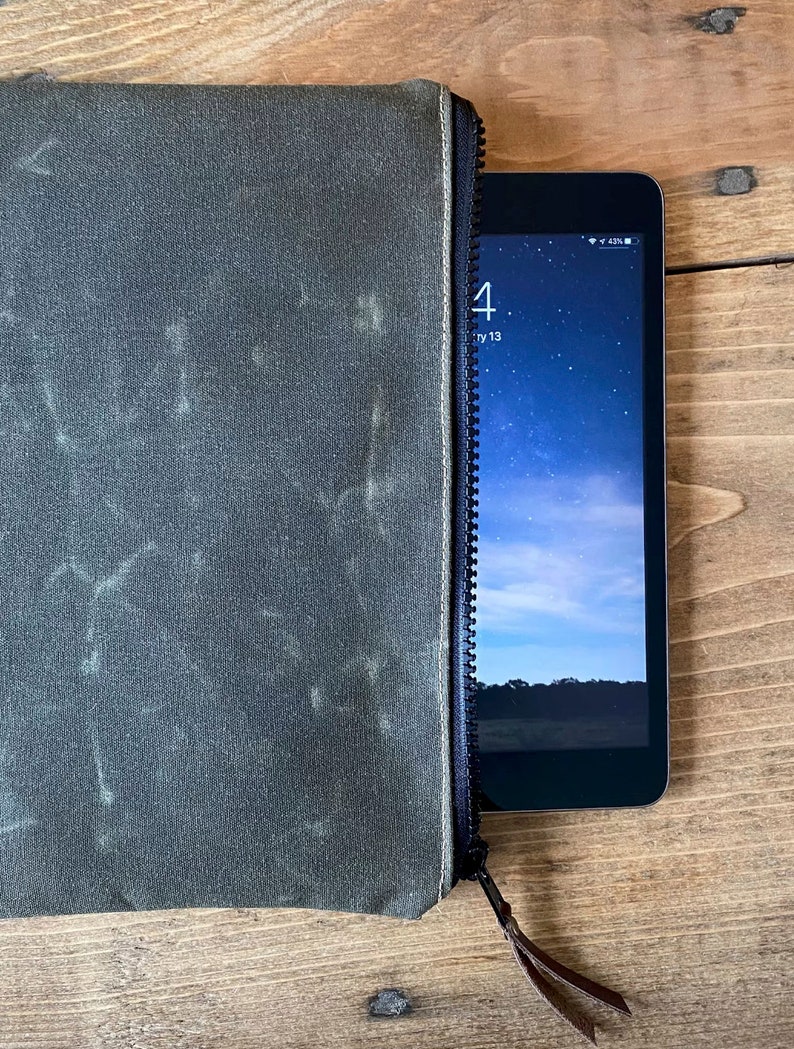 May include: A dark olive green zippered pouch with a textured, weathered finish and a black zipper closure. A small tablet device with a visible screen showing a starry sky background and the time is partially inserted into the open side of the pouch.