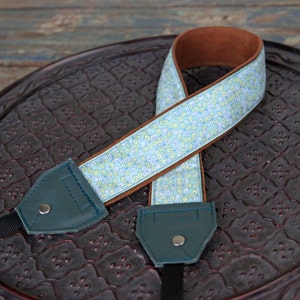 May include: A camera strap with a floral pattern in light blue, yellow, and white. The strap has a brown leather backing and teal leather ends with silver rivets. The strap is designed for a camera.