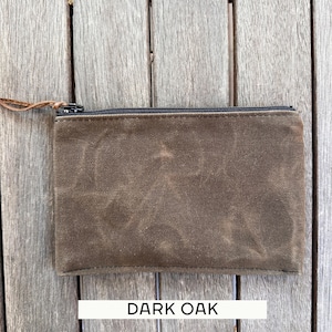 May include: A rectangular dark brown pouch made of a textured, waxed canvas material. The pouch features a black zipper closure along the top edge with a thin brown leather pull tab attached to the metal slider. The text DARK OAK is visible.