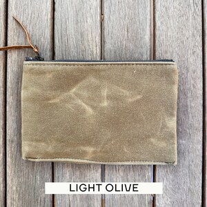 May include: A rectangular light olive zippered pouch made of a textured, waxed-style fabric. The pouch features a black zipper closure along the top edge and a thin, brown leather pull tab attached to the metal zipper slider for easy opening.