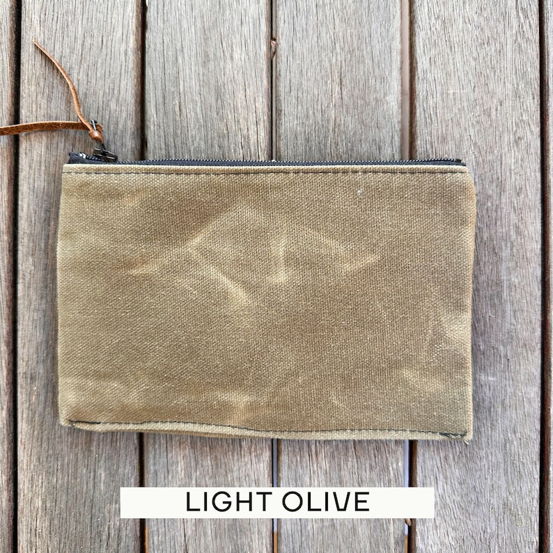 May include: A rectangular light olive zippered pouch made of a textured, waxed-style fabric. The pouch features a black zipper closure along the top edge and a thin, brown leather pull tab attached to the metal zipper slider for easy opening.